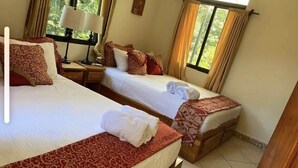 A hotel room with two beds, each with a red patterned bedspread and white linens. There are two bedside tables with lamps, and the room has a window with curtains. - Enchanted Monkey Apartment  (Quepos)
