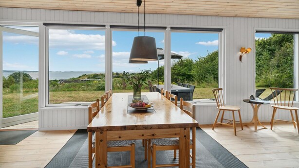 A dining area with a wooden table and chairs, large windows with a view of the sea, and a hanging pendant light. - 3 bedroom beautiful home in Ebeltoft (Ebeltoft)