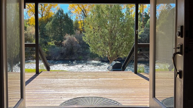 Puerta abierta que da a una terraza de madera con vistas a un río y árboles.
