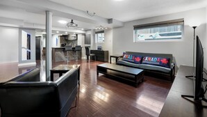 A spacious living area with a sofa, coffee table, and a television. A kitchen is visible in the background, and there is a window with a view of a snowy landscape. - Stylish 1 bedroom basement unit with renovated bathroom & in-suit Jacuzzi. (Richmond Hill)
