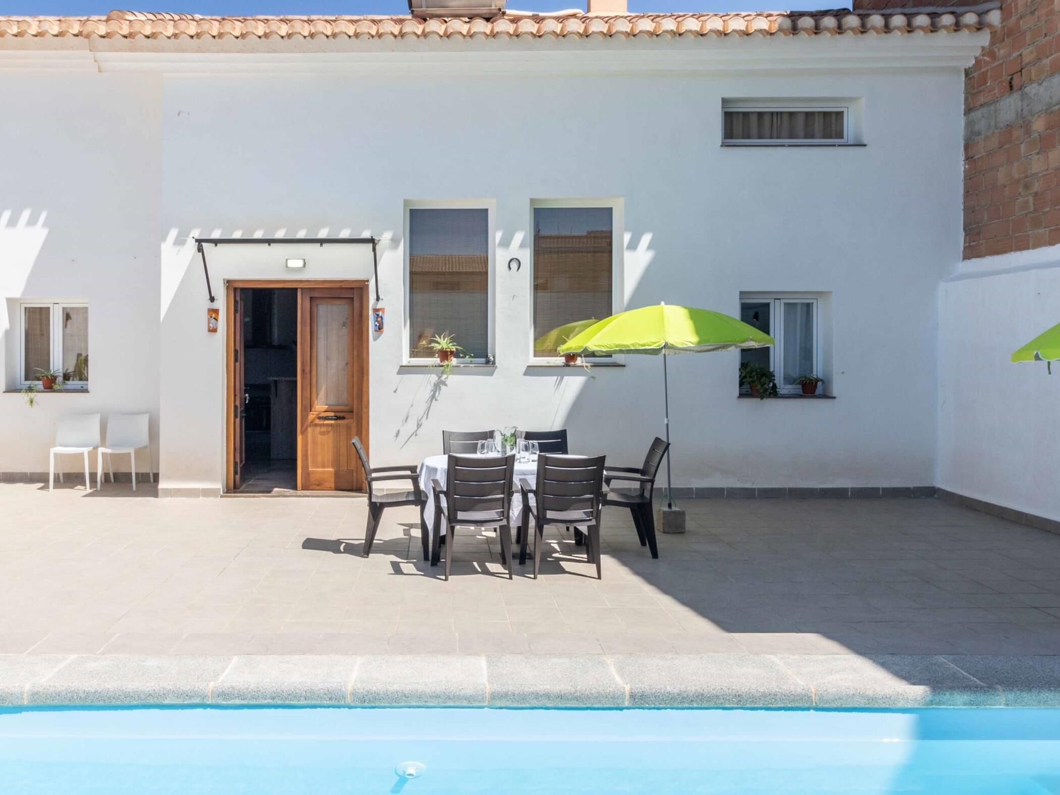Un espace aménagé au bord de la piscine, avec une table et des chaises, une porte en bois et une fenêtre ornée d’une plante.