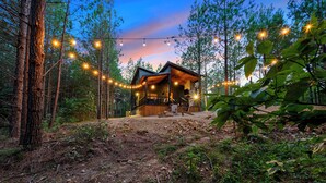 Una cabaña en un bosque, con luces colgantes, rodeada de árboles y un sendero de tierra.