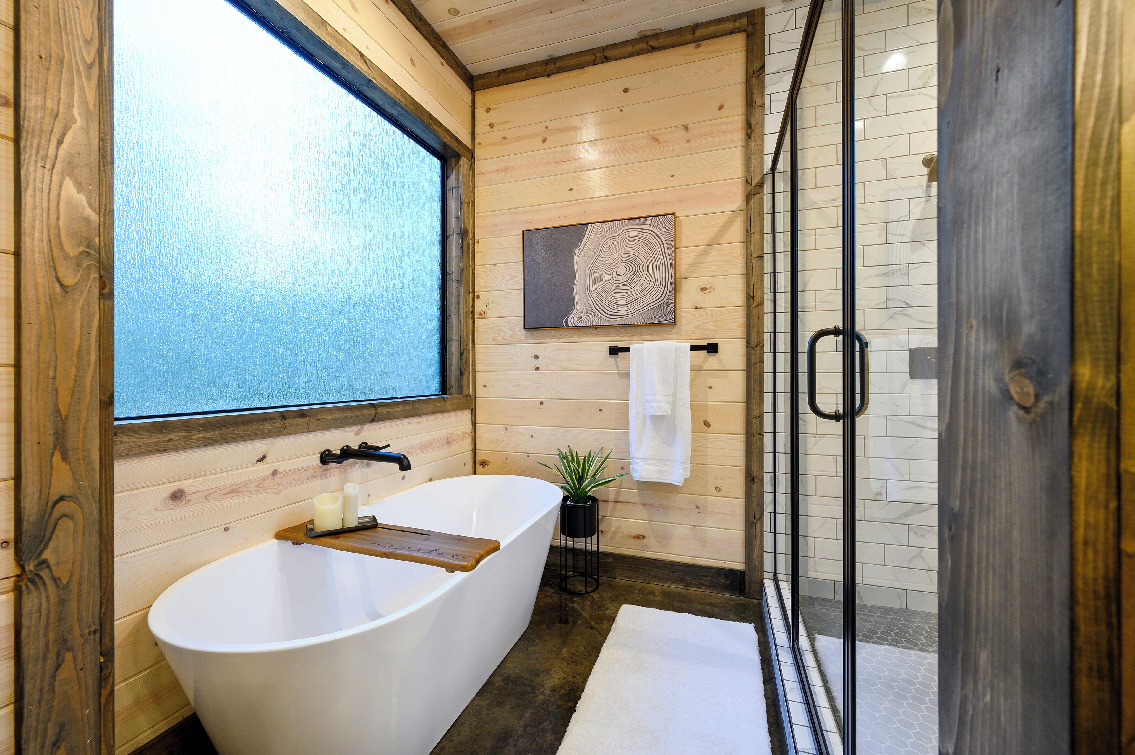 A modern bathroom with a freestanding bathtub, a glass shower enclosure, wooden walls, and a large window providing a view.