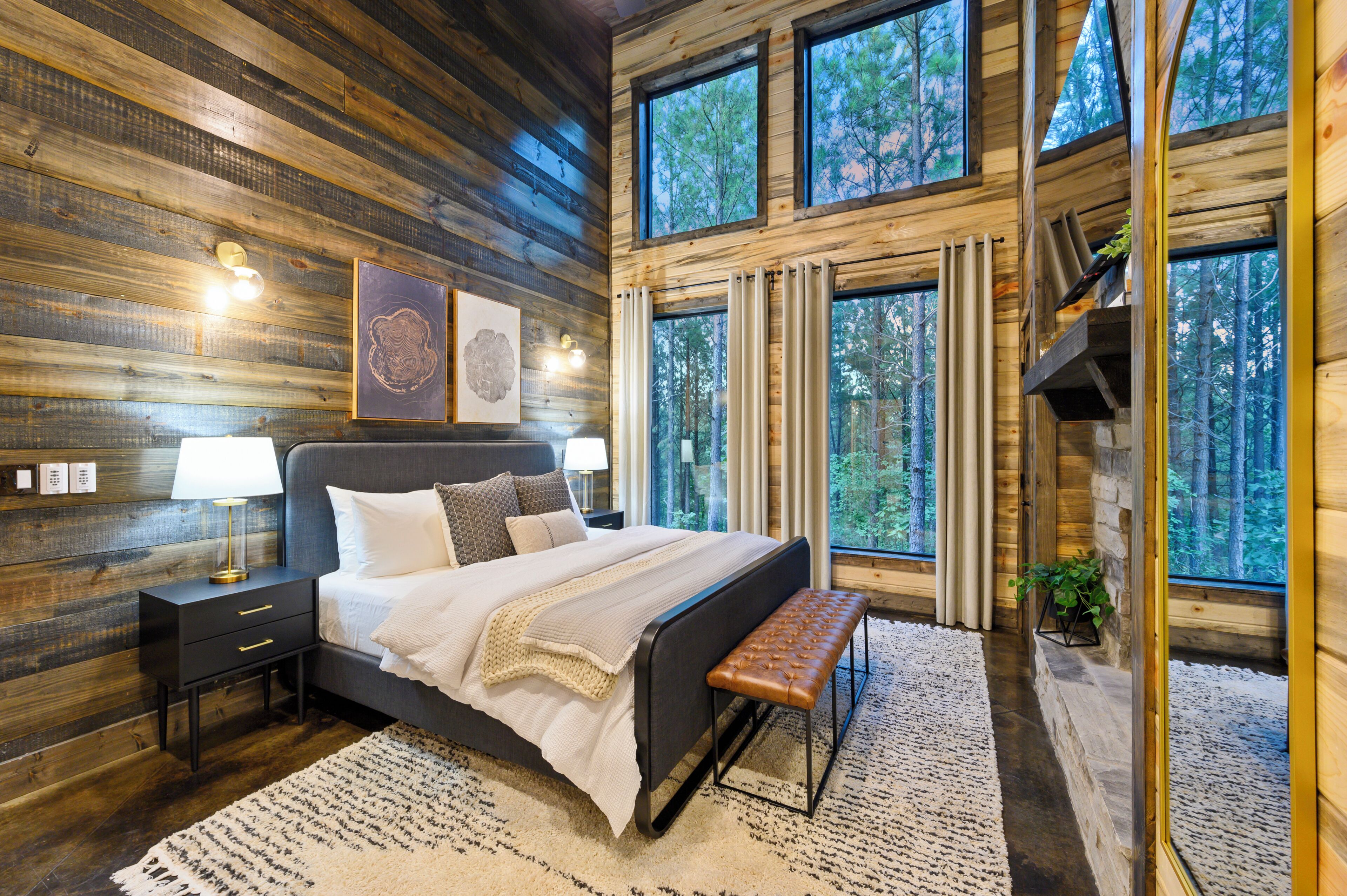 A bedroom with a large bed, bedside tables, a bench, and a view of the forest through large windows.