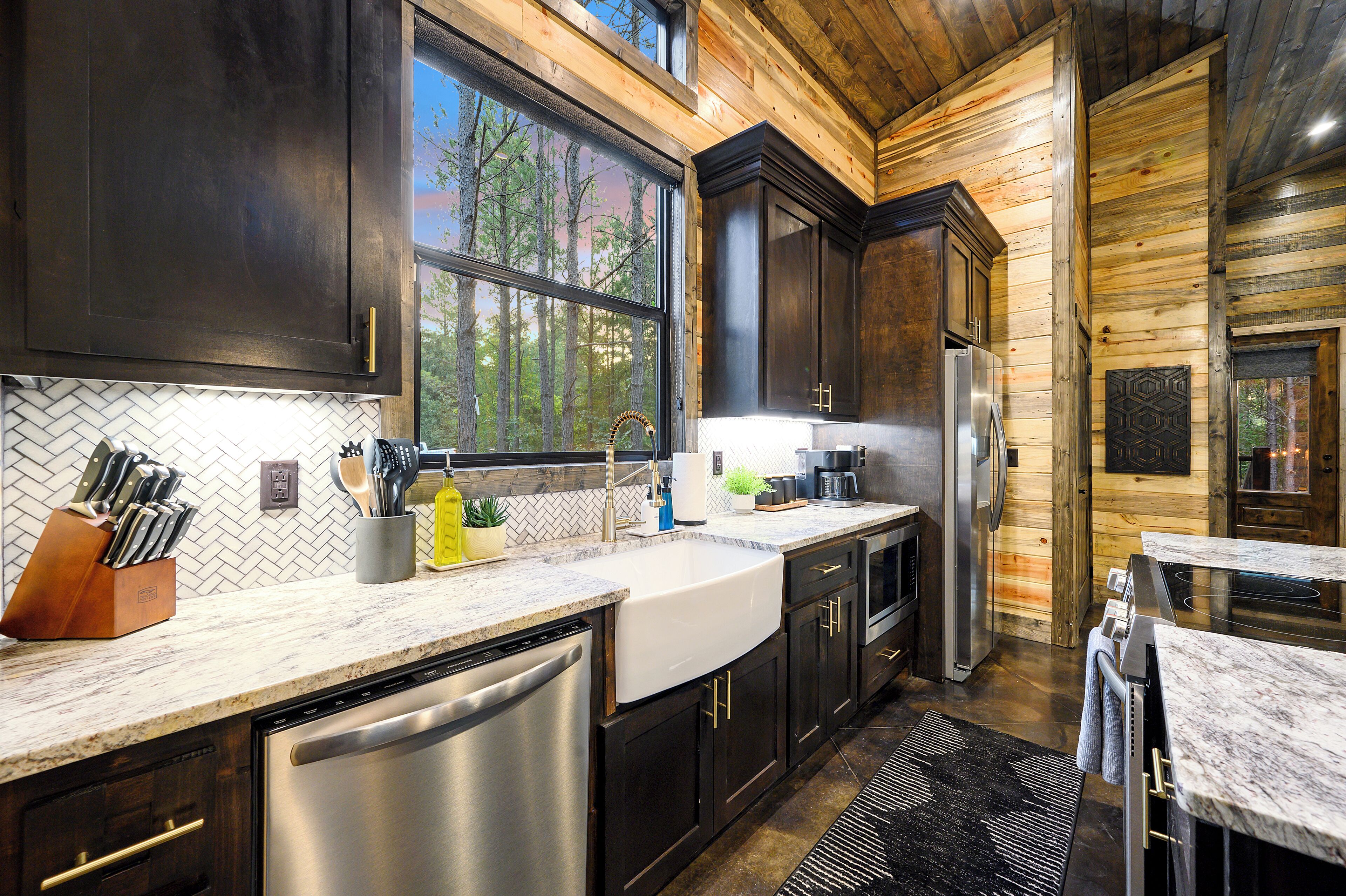 A modern kitchen with dark wood cabinets, stainless steel appliances, and a large window offering a view of trees.