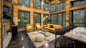 Amplia sala de estar con un ventanal que da a un bosque, una chimenea y una mesa de comedor con sillas.