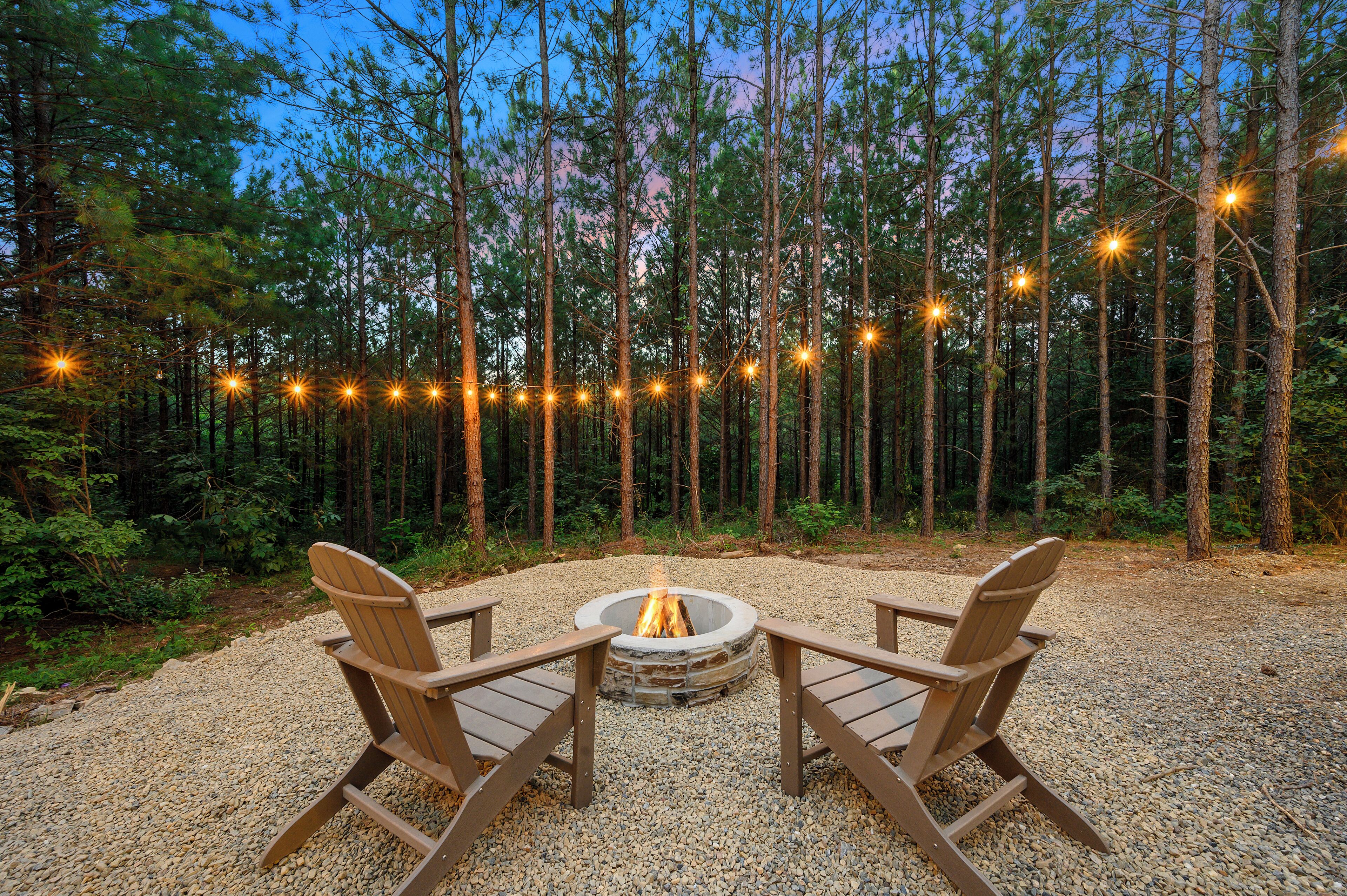 Un foyer circulaire, entouré d’une zone de gravier et bordé d’une forêt, avec deux fauteuils Adirondack en bois placés face à lui.