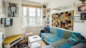 Ein gemütliches Wohnzimmer mit einer Couch, einem kleinen Tisch, einem Sessel, einem Bücherregal und einer großen Stegdecke an der Wand.