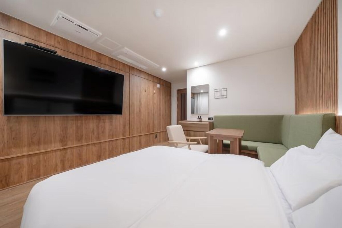 Une chambre d’hôtel moderne avec un grand lit, un téléviseur à écran plat fixé au mur, une armoire en bois, un petit bureau et un canapé vert.