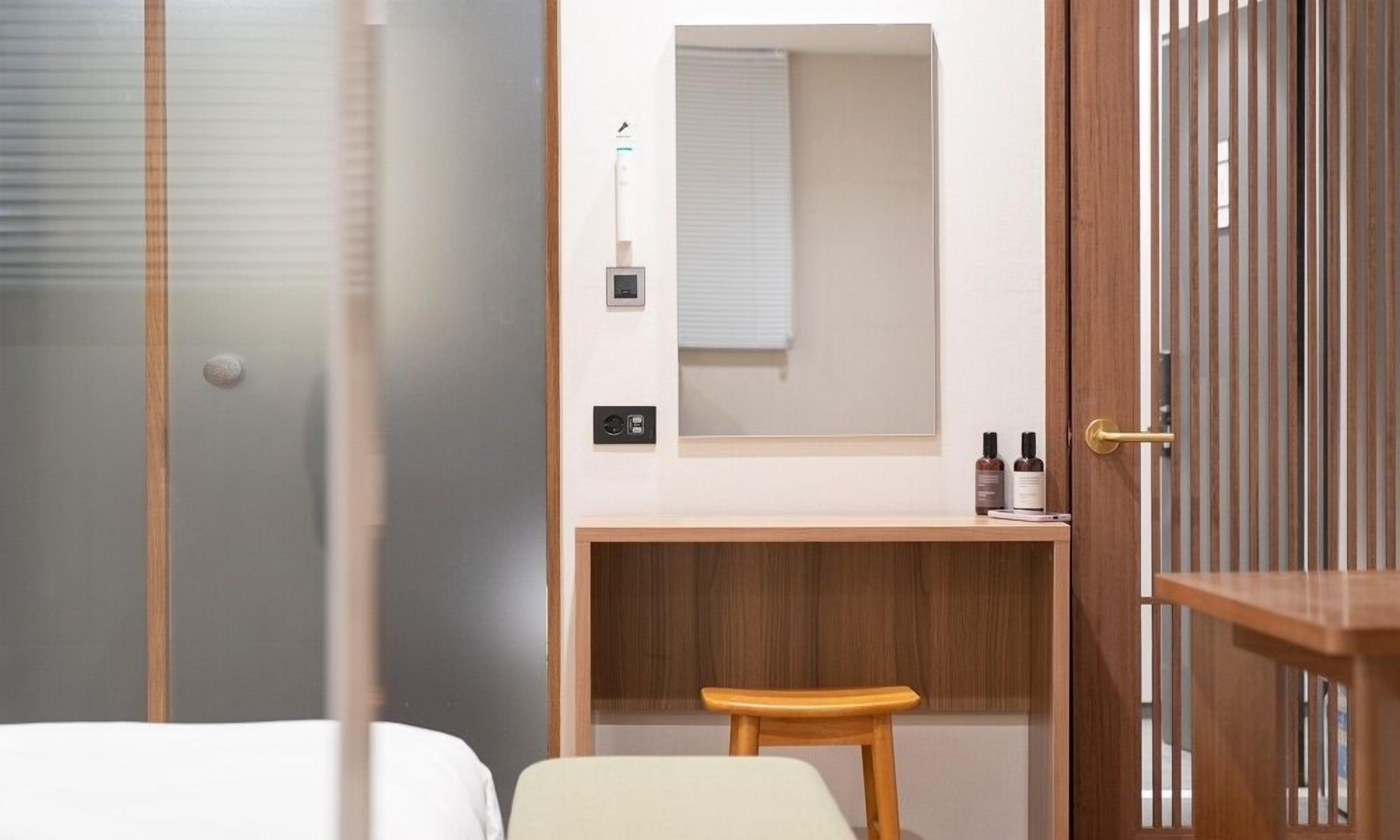 A hotel room with a bed, a wooden desk, a mirror, and a small stool.