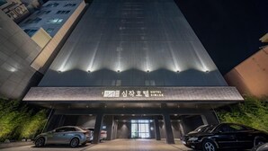 A modern hotel entrance with a reflective facade, a sign indicating it is the 'SIMJAK HOTEL', and a parking area with cars. - Simjak Hotel Dongtan By Anook (Hwaseong)