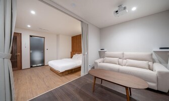 Una habitación de hotel moderna con suelo de madera, una cama blanca, un sofá color beige y una pequeña mesa de madera.