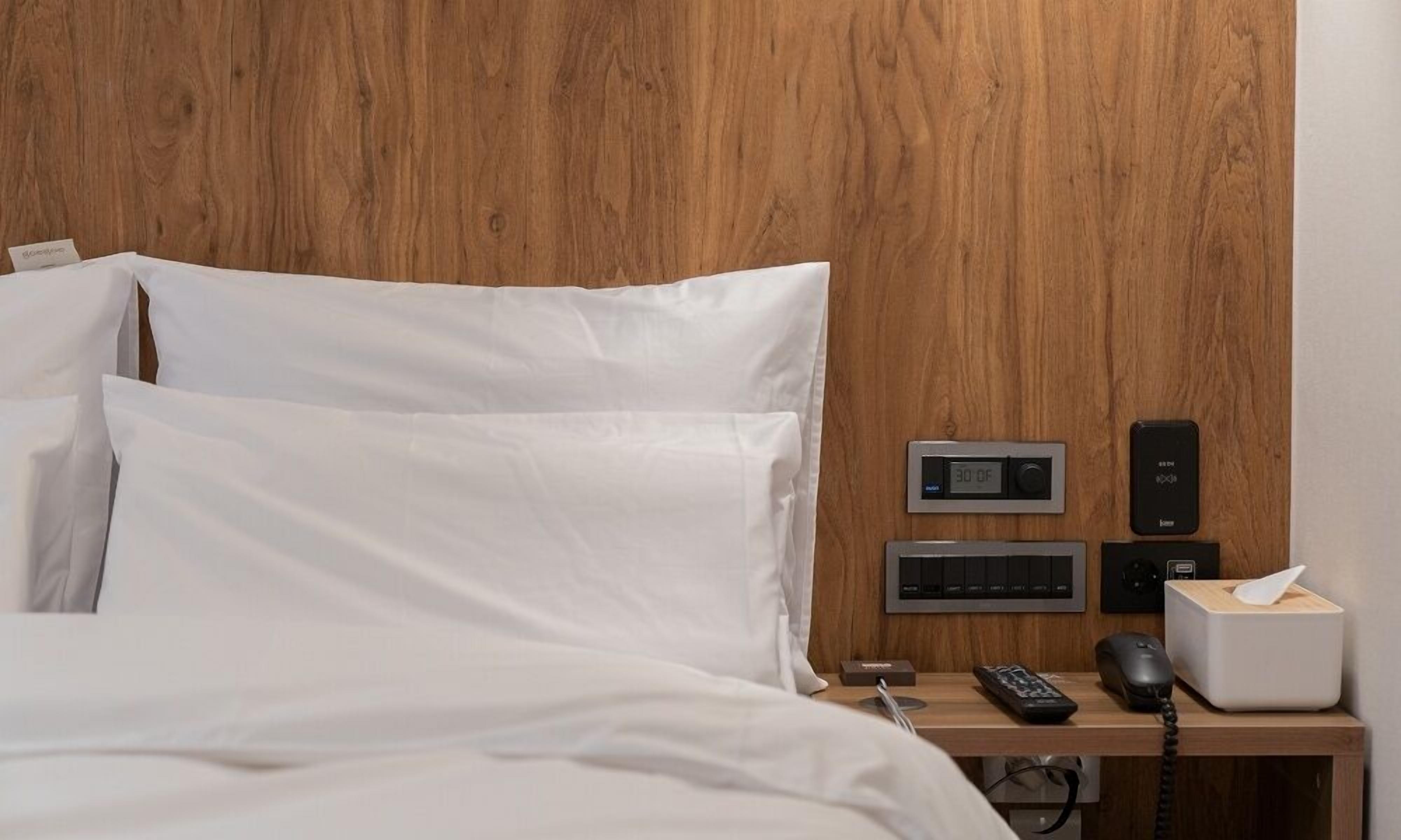 A neatly made bed with white linens, a bedside table with a telephone, a digital clock, a remote control, and a tissue box.