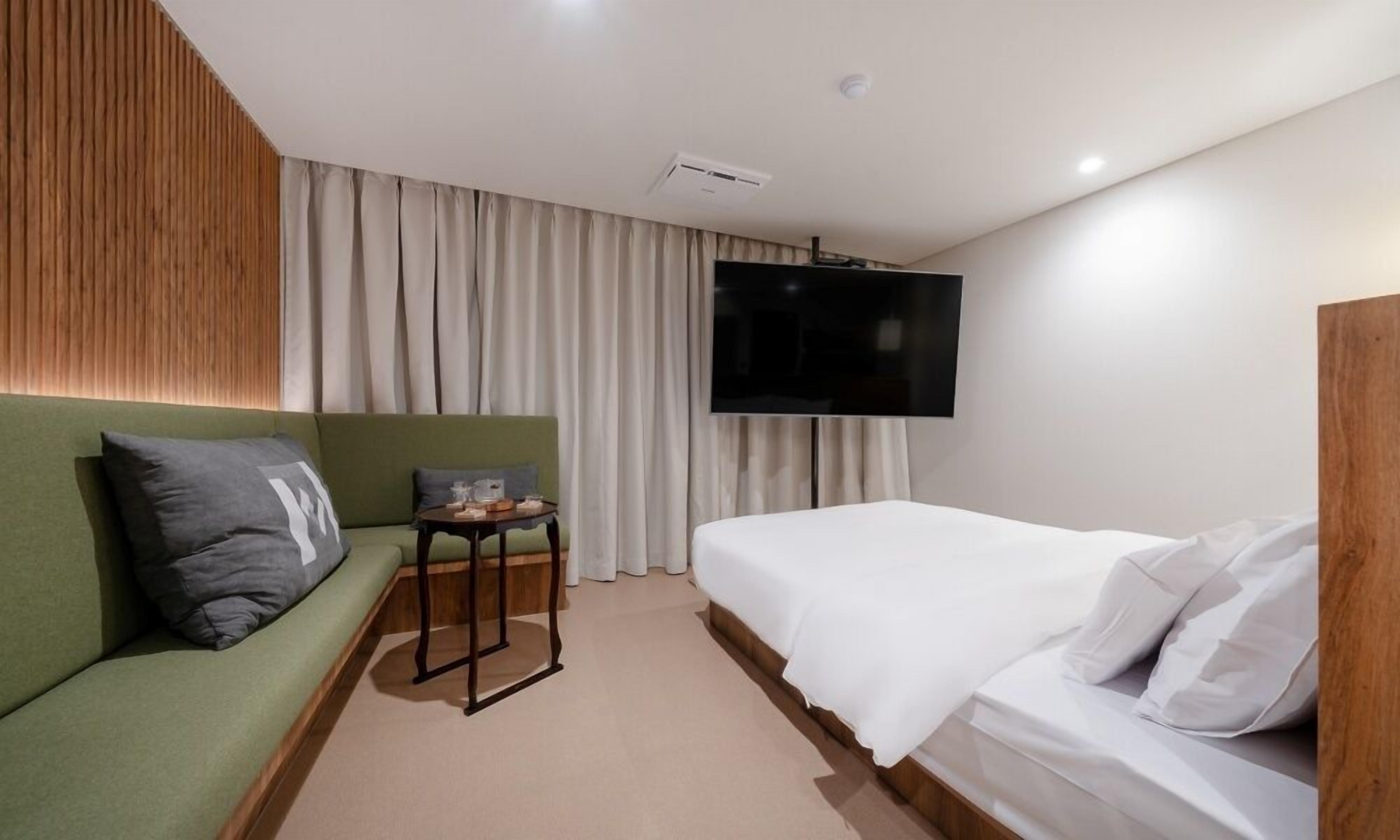 Habitación de hotel con un sofá verde, una pequeña mesa, una cama con ropa de cama blanca y un televisor montado en la pared.