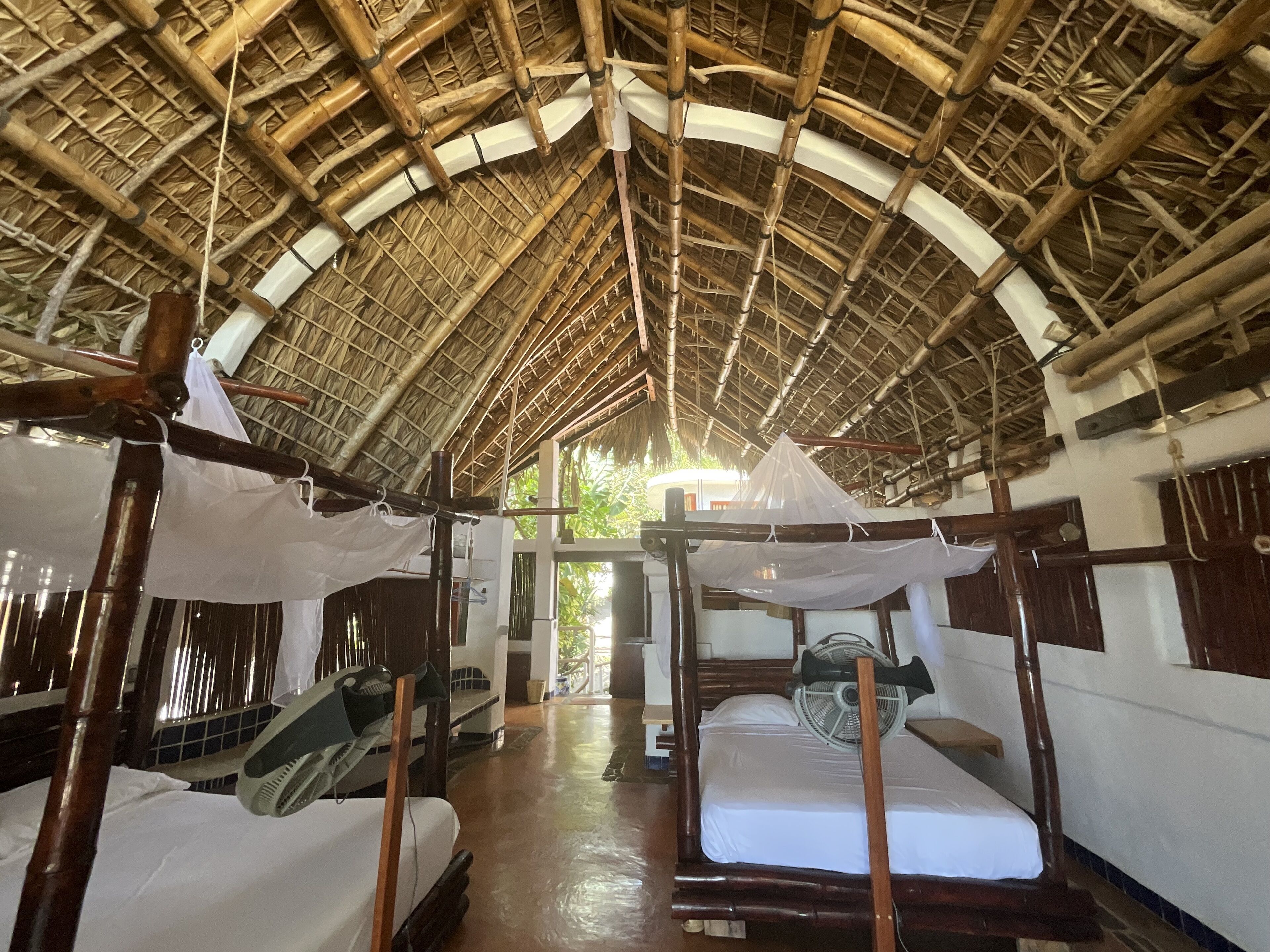 A room with a thatched roof, wooden beds, and a fan.