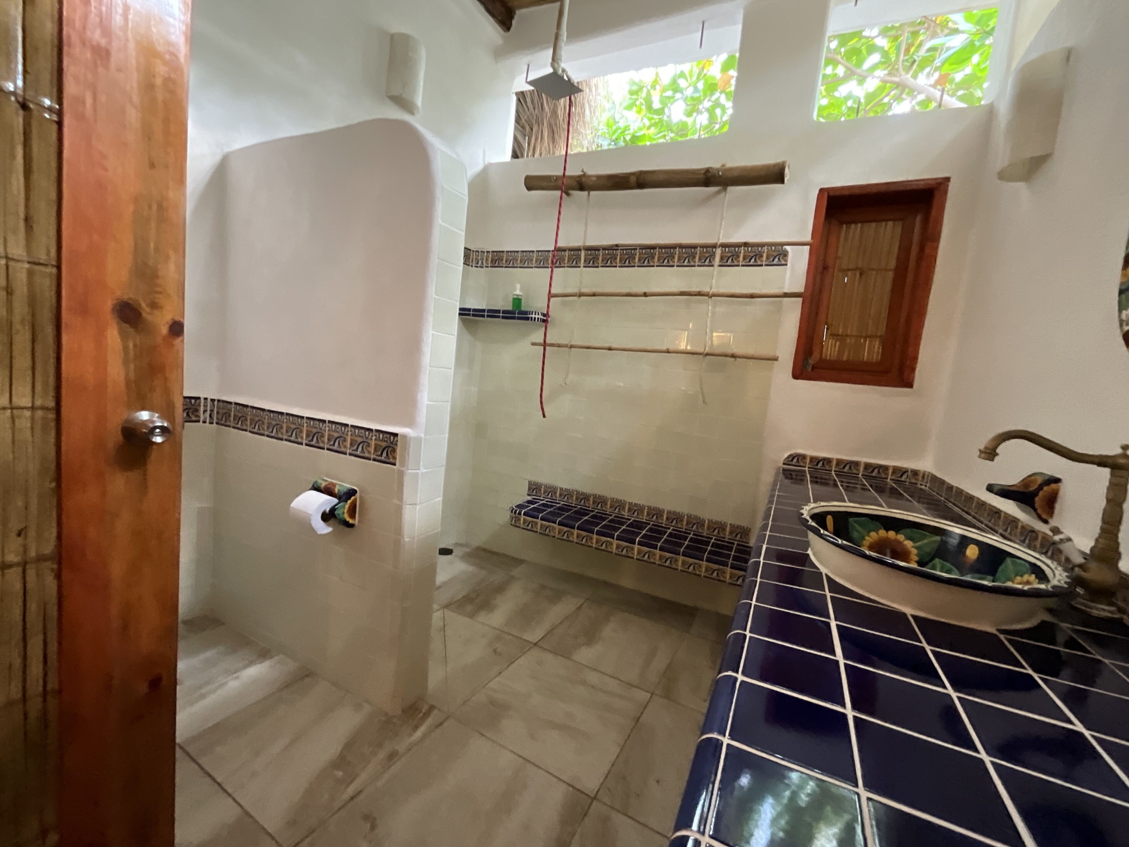 A bathroom with a tiled floor, a wooden door, a towel rack, a shelf with toiletries, and a decorative basin.