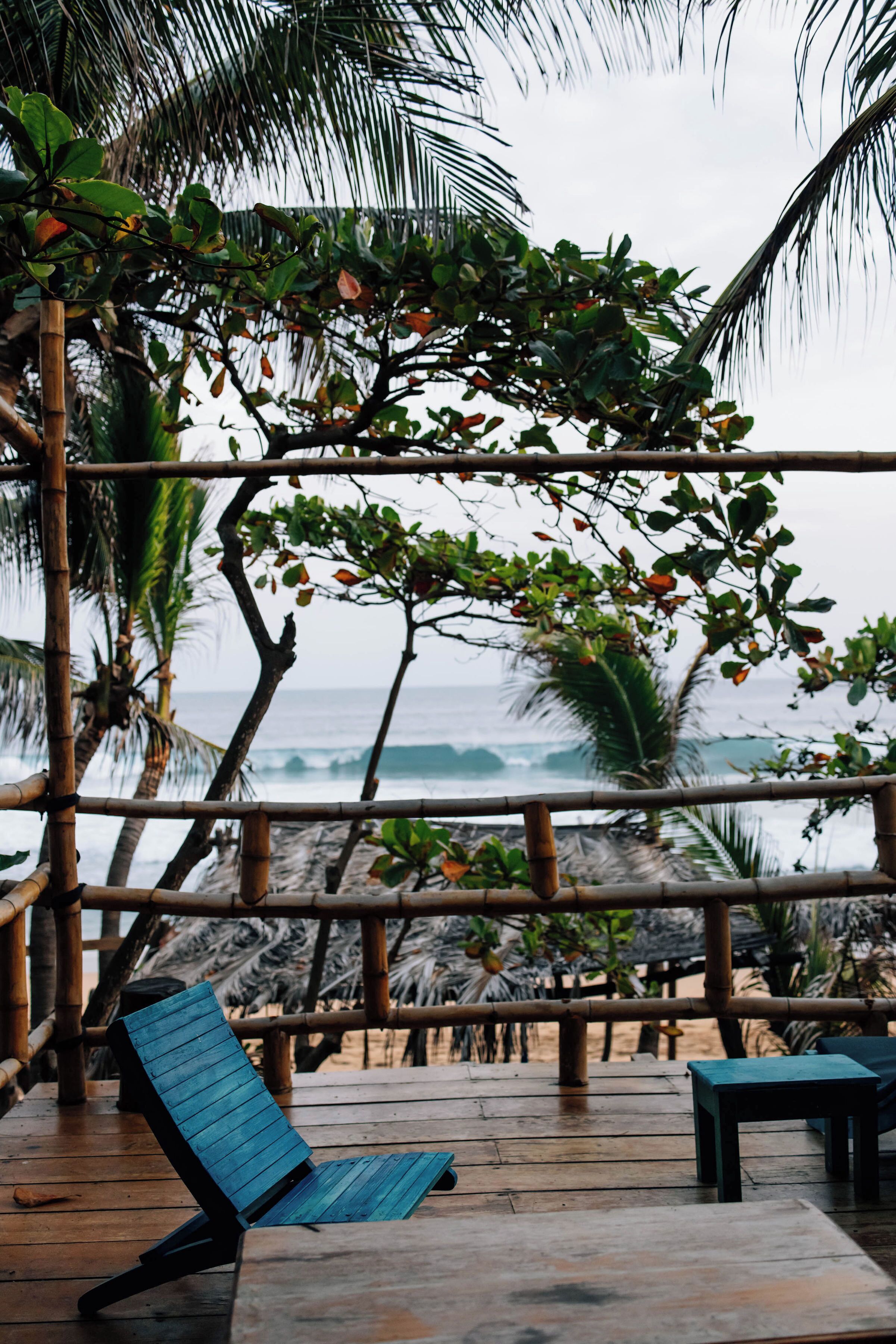 Une terrasse en bois avec une chaise bleue et une table, d’où l’on a une vue sur l’océan, avec des palmiers et des structures en bambou.