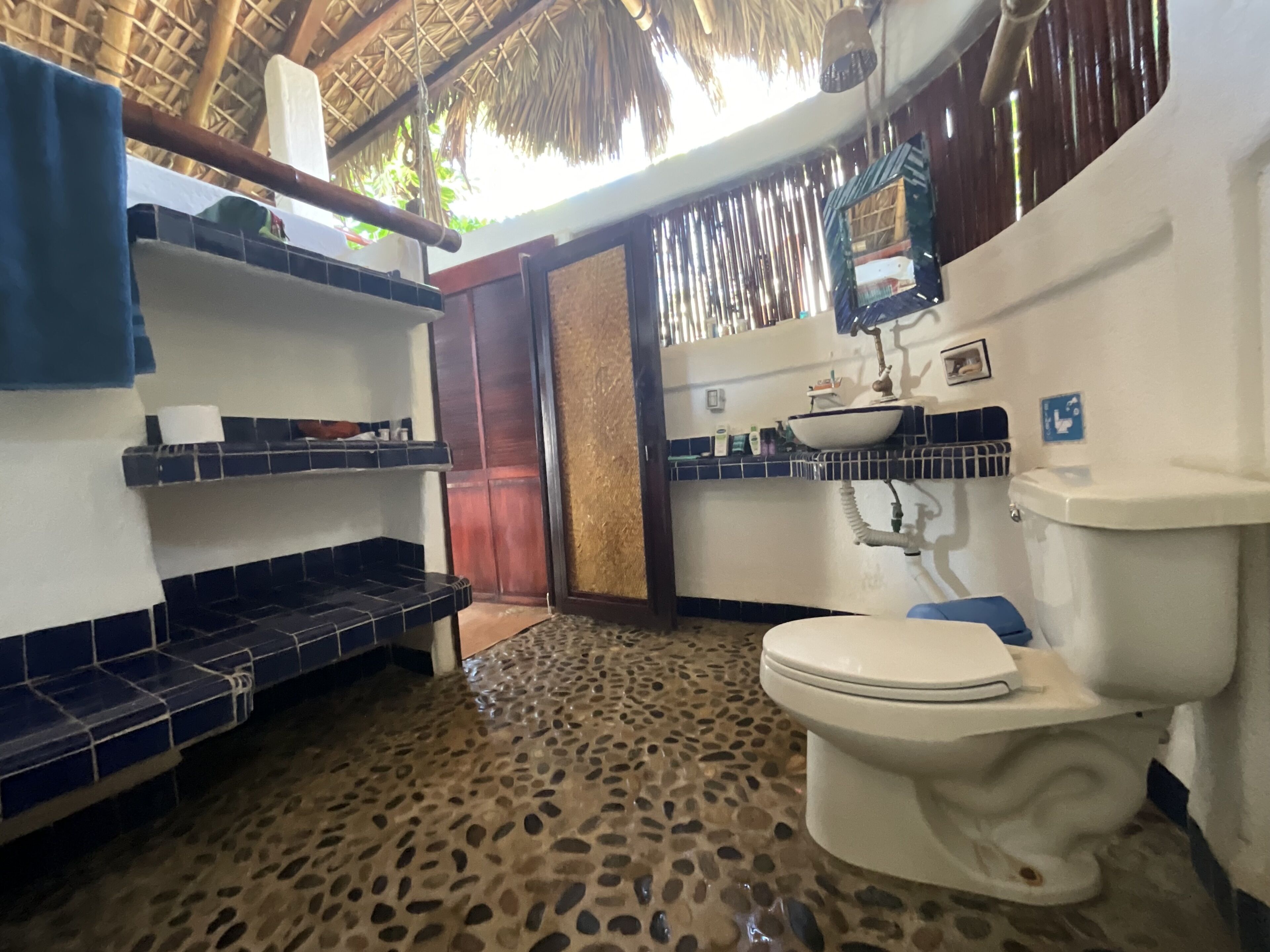 Un baño con suelo de baldosas, inodoro, lavabo y estantes con artículos de aseo.