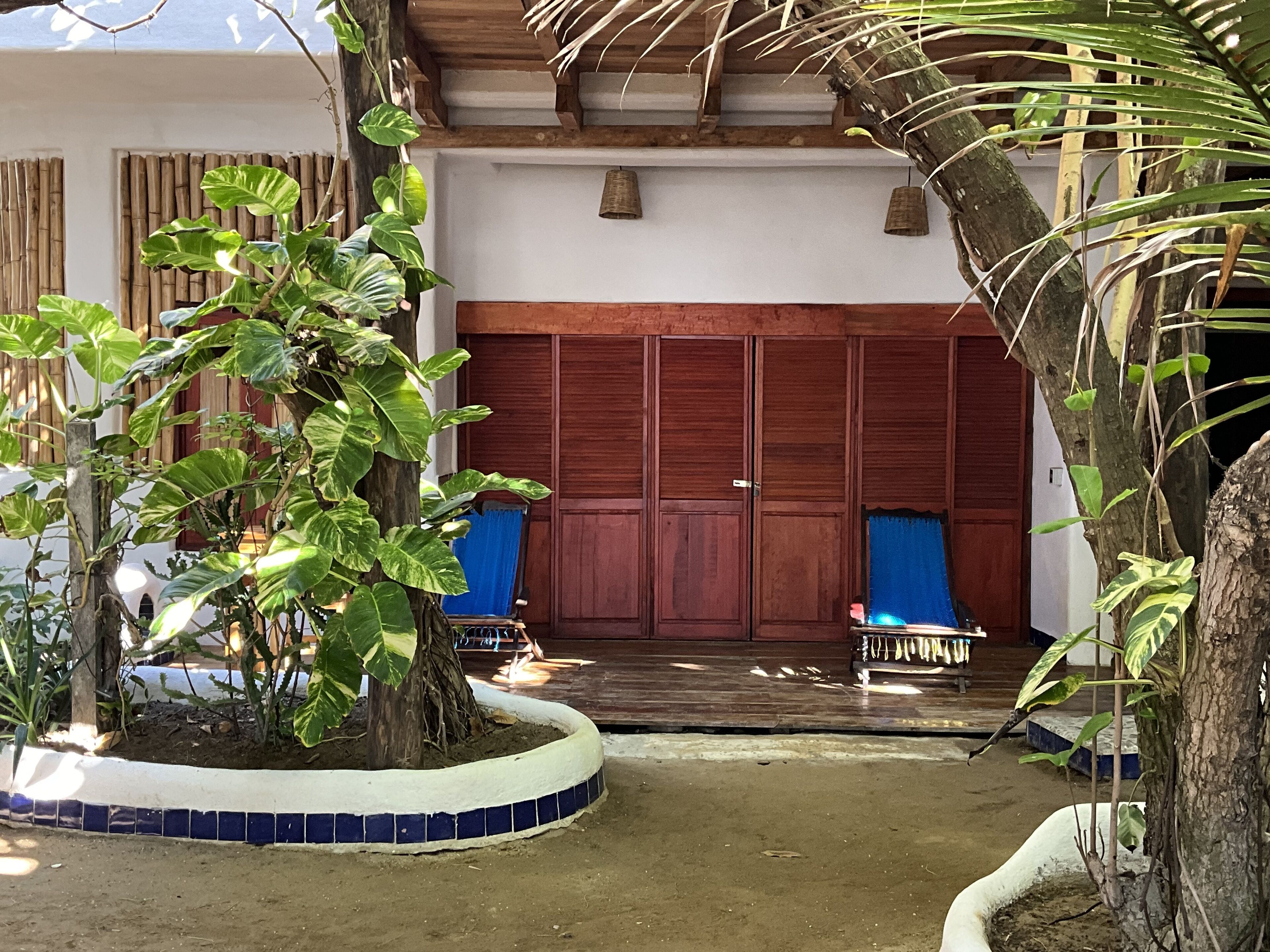 A courtyard with a wooden door, a tiled floor, and a variety of plants.