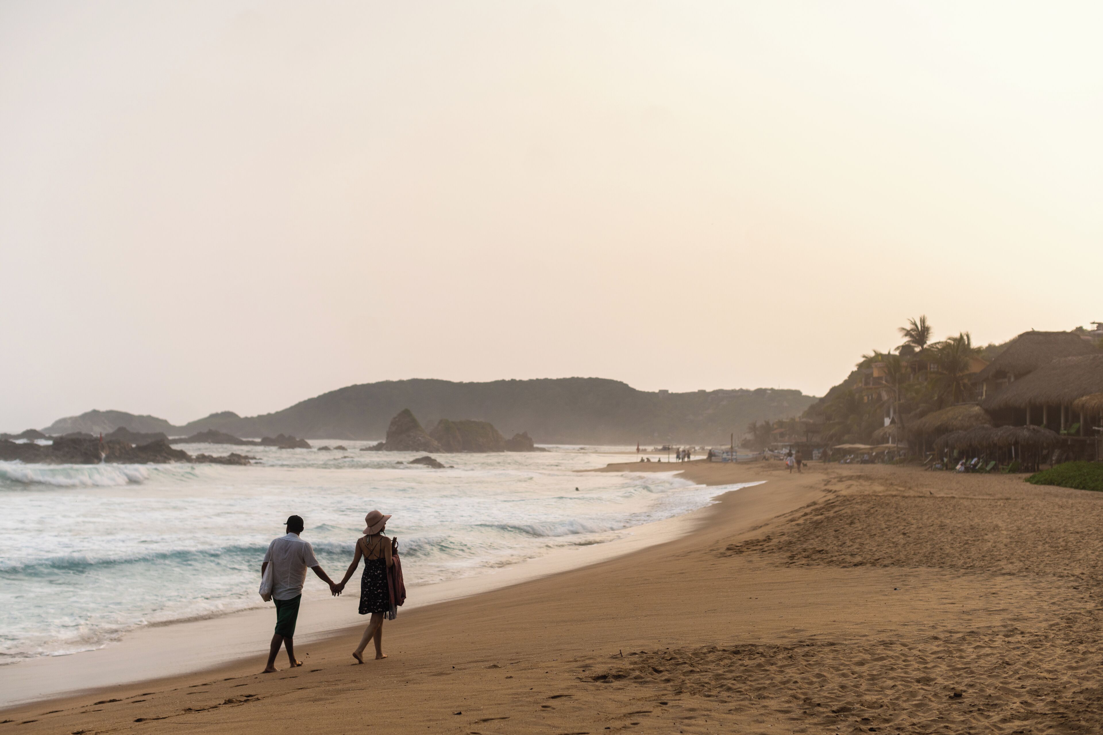 Un couple se promène sur une plage de sable, avec des vagues à l’arrière-plan et un paysage vallonné.