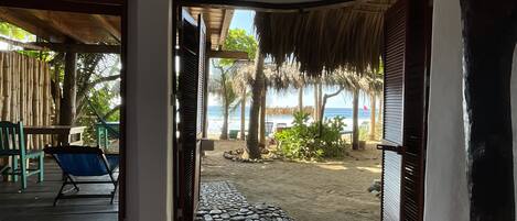 Blick auf einen Strand von einer überdachten Terrasse mit Tisch und Stühlen, einem Reetdach und einem Steinpfad, der zum Strand führt.