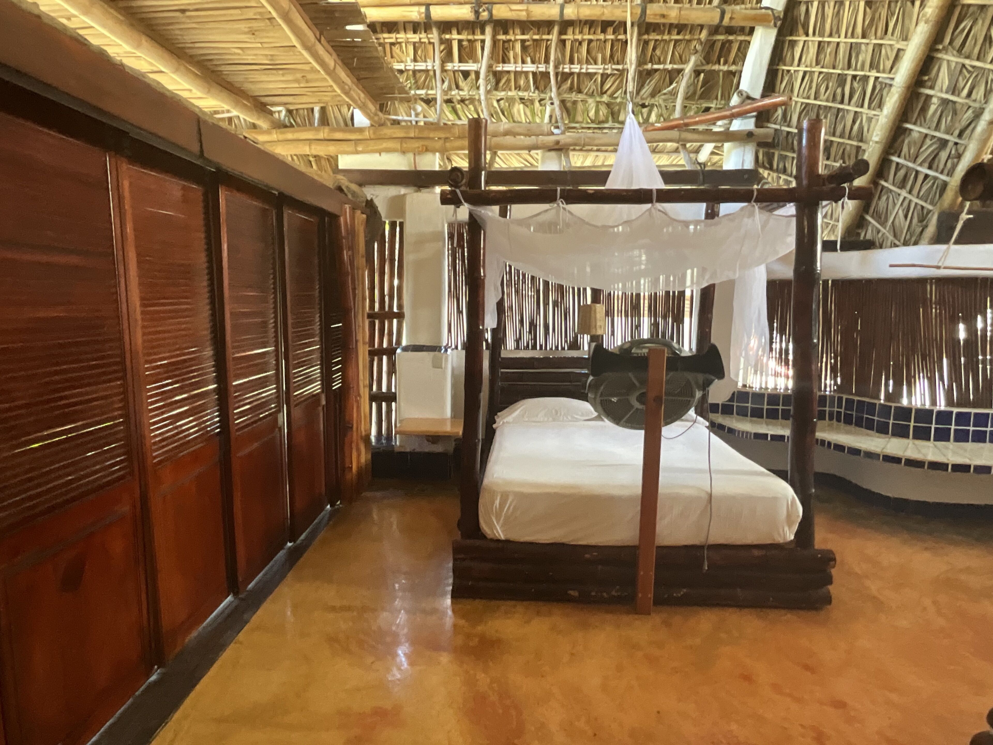 A traditional room with a thatched roof, a wooden bed with a mosquito net, and a wooden wardrobe.