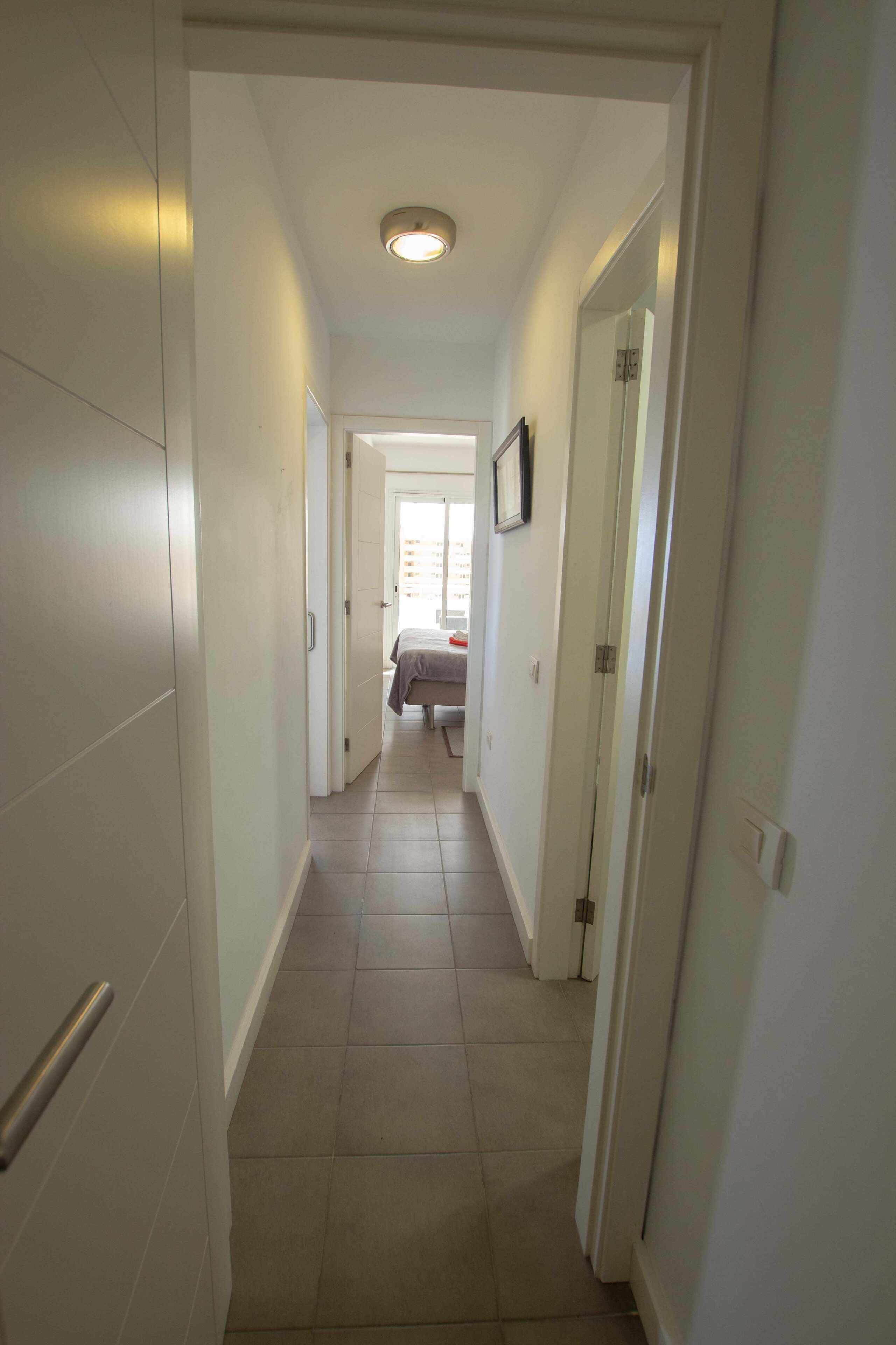 A narrow hallway with white walls and a tiled floor leading to a bedroom with a bed and a window.