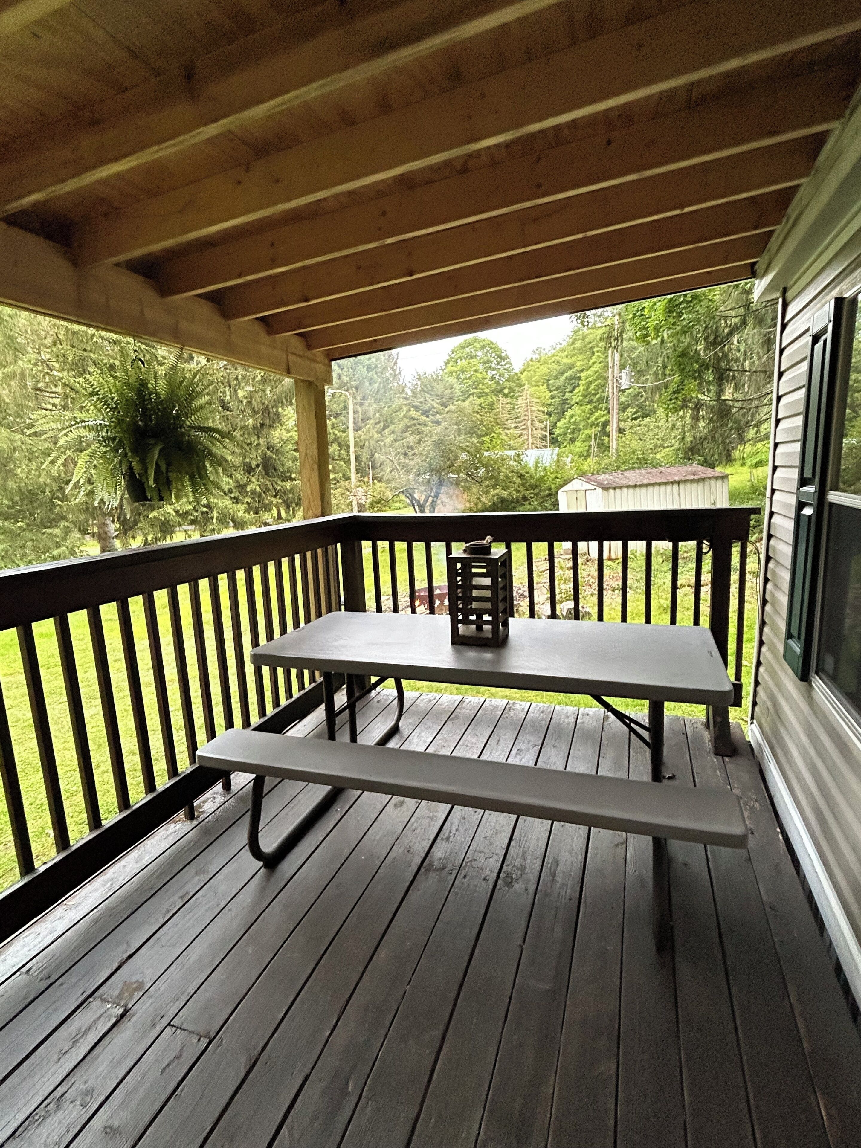 Un espace extérieur abrité, doté d’une table de pique-nique et de bancs, entouré d’une rambarde en bois, et offrant une vue sur une pelouse et des arbres.