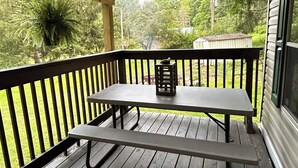 A covered outdoor area with a picnic table and benches, surrounded by a wooden railing, overlooking a grassy yard and trees.