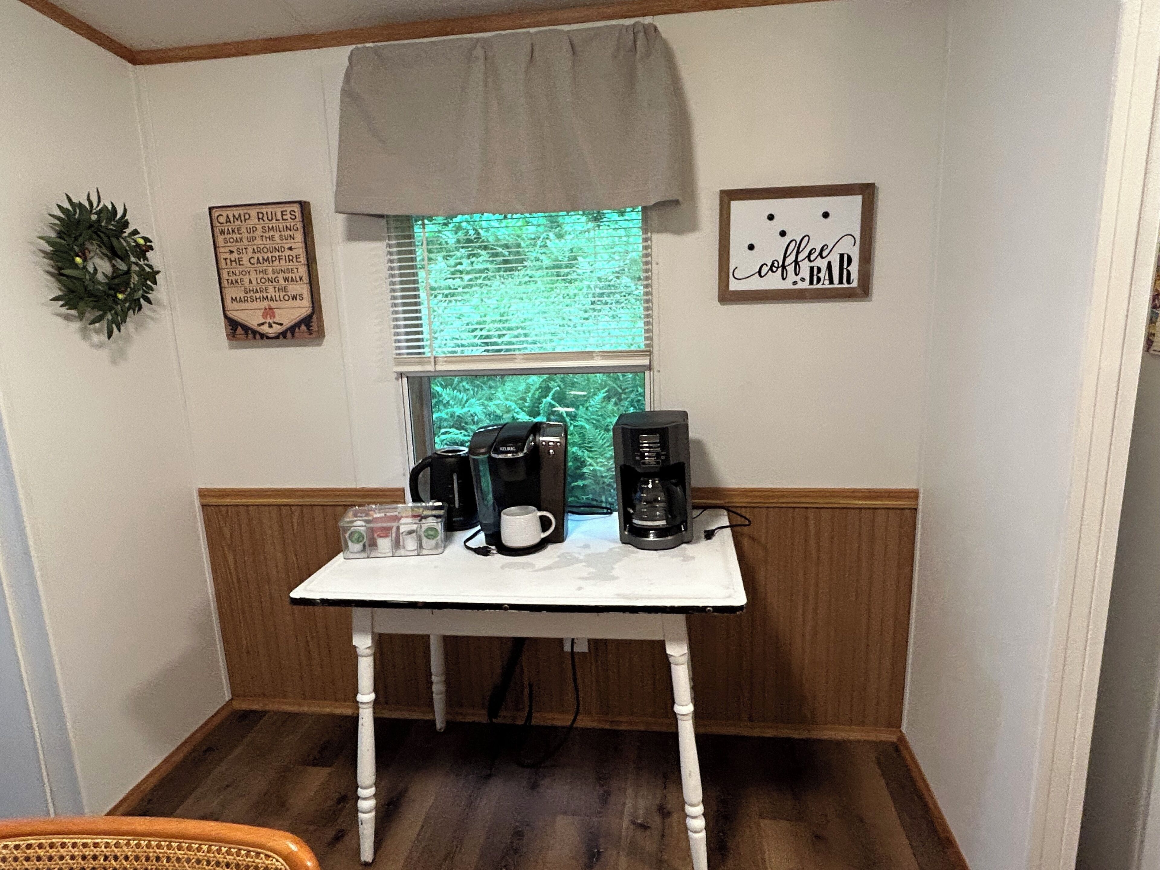 Une petite table blanche, sur laquelle se trouvent une machine à café, une tasse et des récipients, est placée dans un coin orné de lambris en bois et près d’une fenêtre munie de stores.