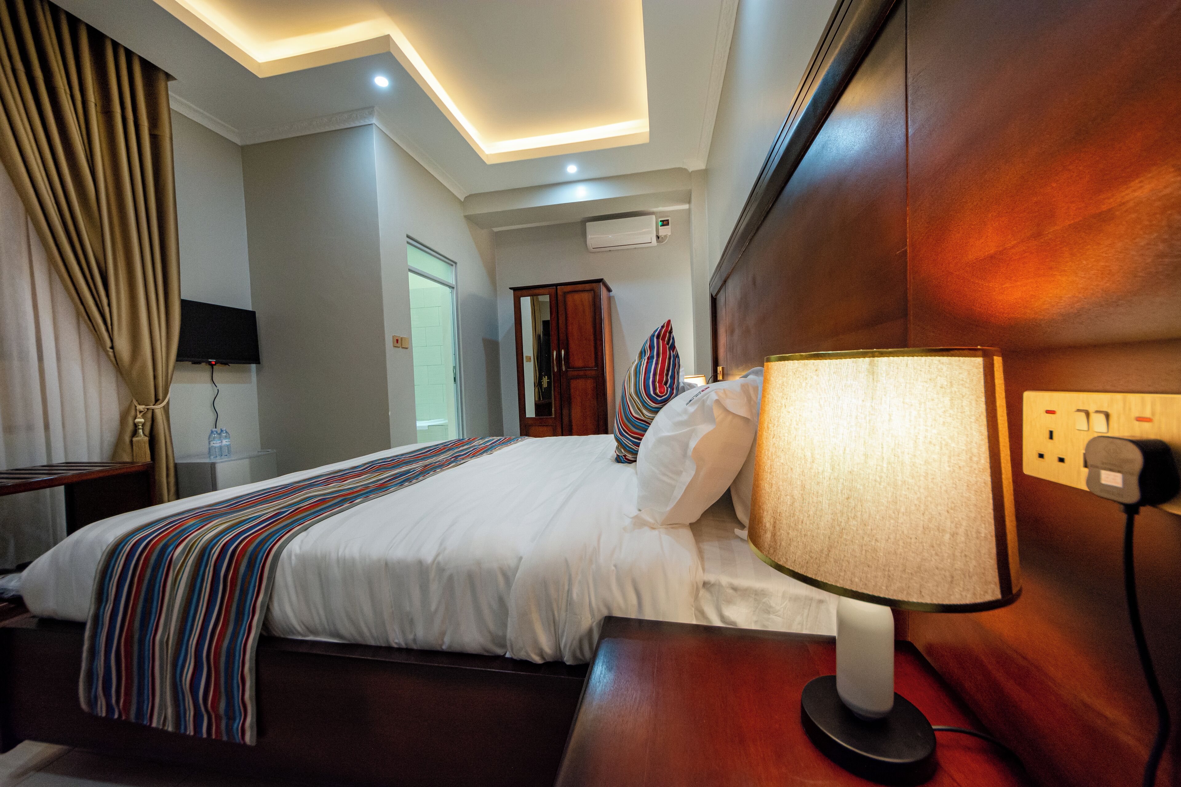 A hotel room with a large bed, a bedside lamp, a flat-screen TV mounted on the wall, and an air conditioning unit.