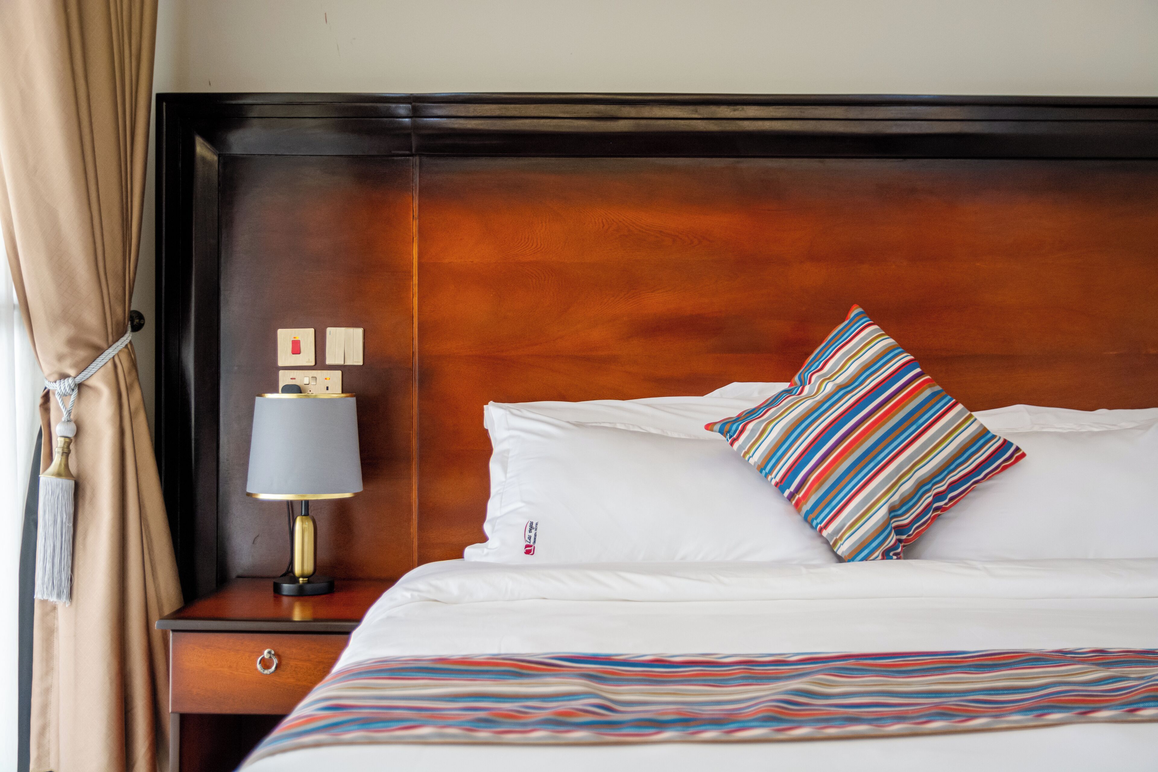 A neatly made bed with white linens and striped pillows, a bedside lamp, and a wooden headboard.