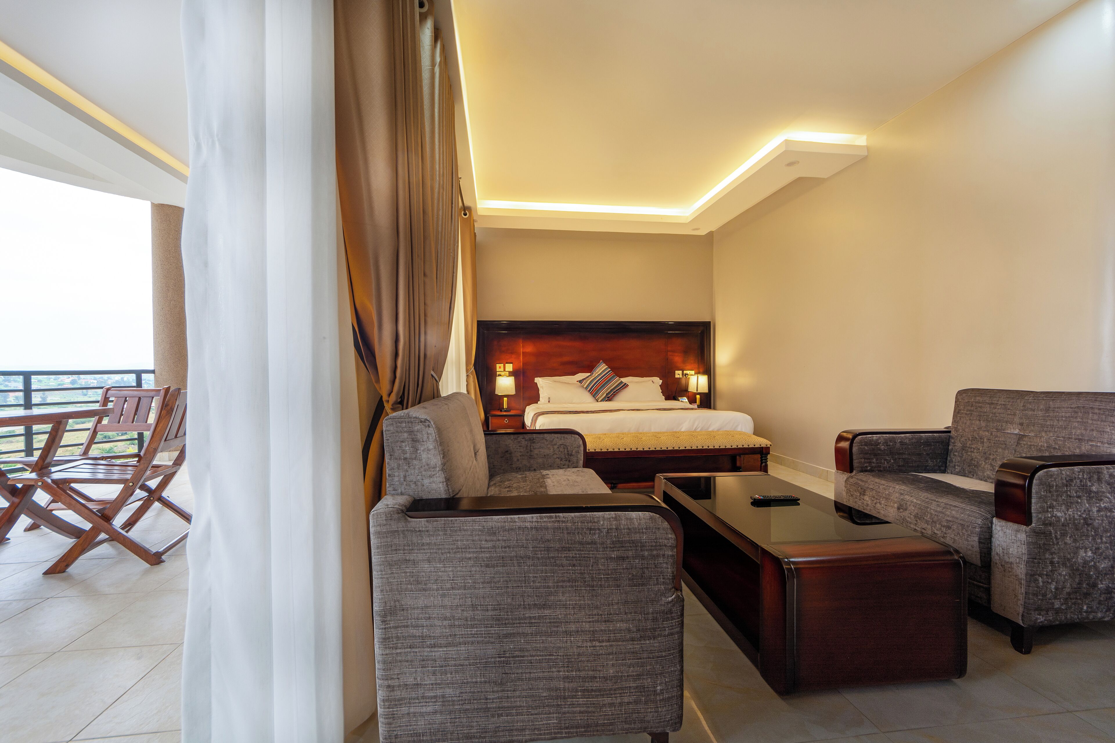 A hotel room with a bed, two armchairs, a coffee table, and a balcony with wooden chairs.