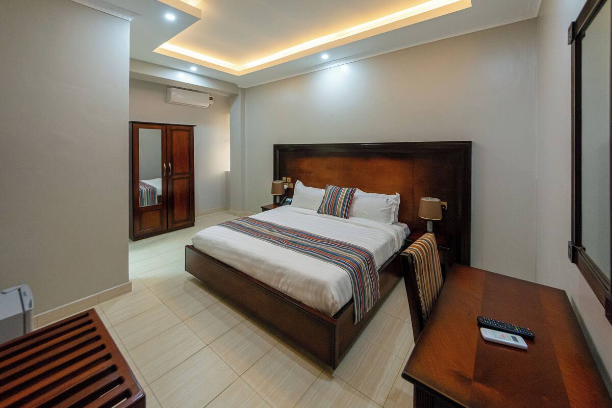 Una habitación de hotel moderna con una cama grande, mesitas de noche, cabecera de madera y una pequeña mesa con un control remoto.