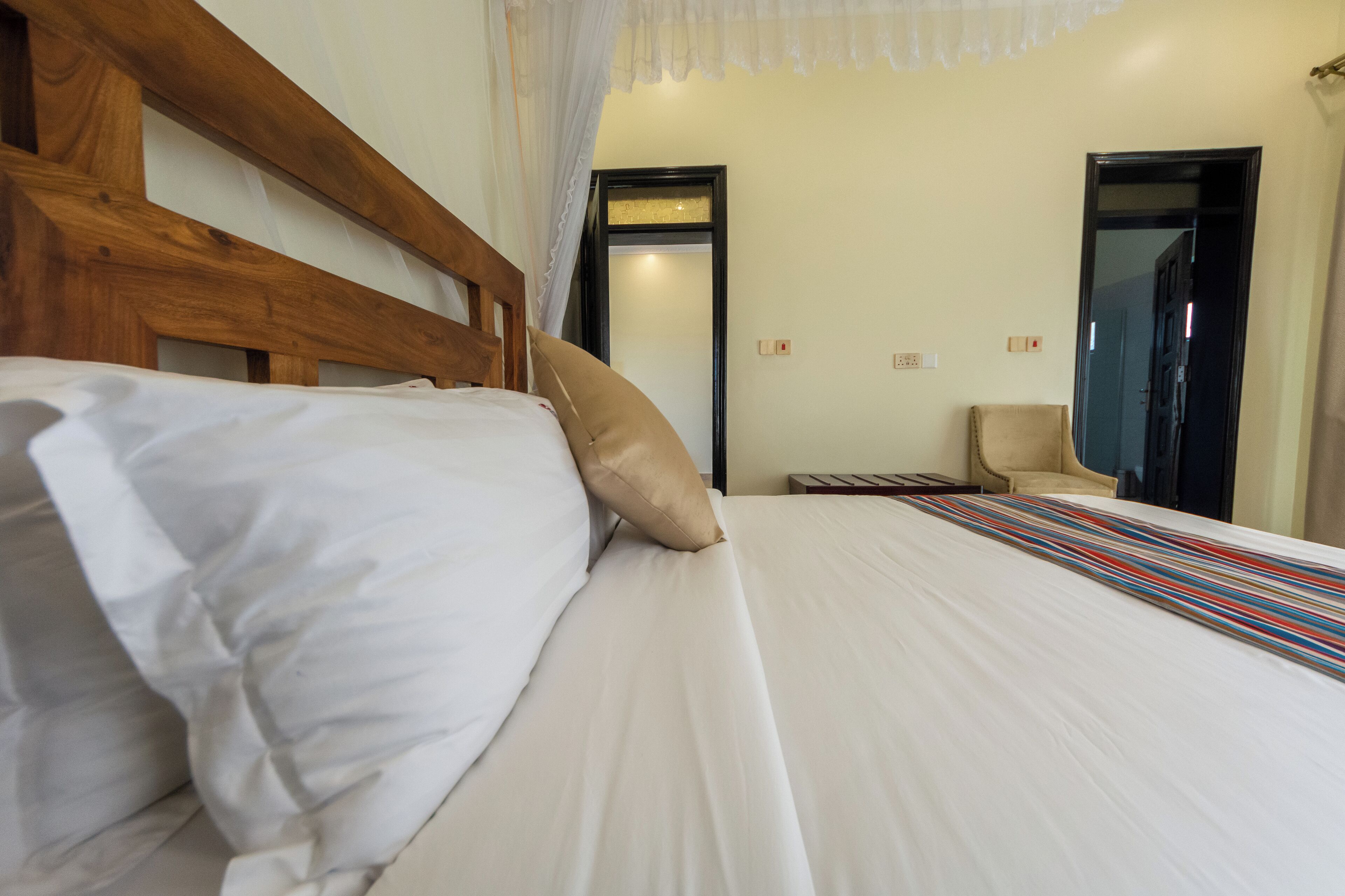 A neatly made bed with white linens and a striped bedspread, a wooden headboard, and a chair in the background.