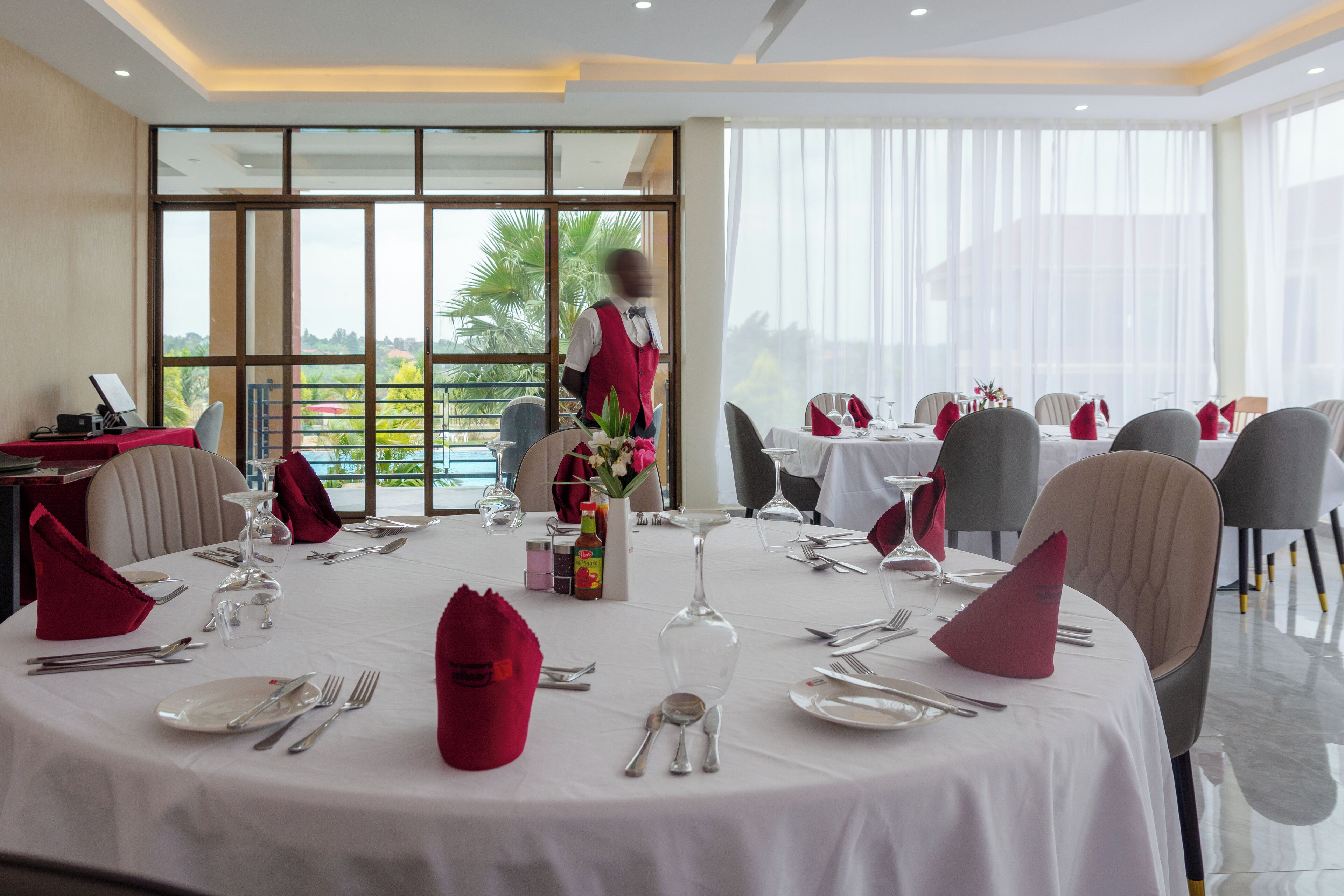 A restaurant with round tables set for dining, large windows with a view, and a waiter in uniform.