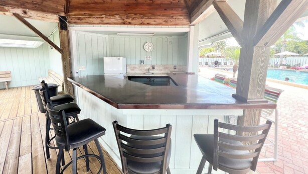 Cocina exterior techada con barra, terraza de madera y vista a una piscina.
