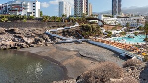A coastal resort with a rocky beach, a swimming pool, and high-rise hotels in the background. - Cozy Apartment with Sea Views in Costa Adeje (Adeje)
