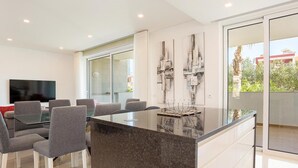 A modern kitchen with a central island, a dining area with grey chairs, and a large window offering a view of greenery. - Luxury Apt. Garden, Pool and Near Beach (Lagos)