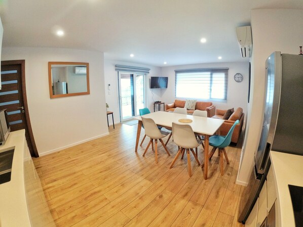 A spacious living area with a wooden floor, a dining table with chairs, a sofa set, a TV mounted on the wall, and a refrigerator. - 40 Warren Street (Wanaka)