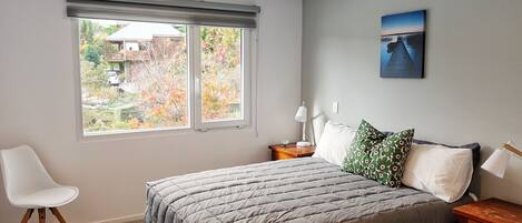 Ein Schlafzimmer mit Bett, Sessel, Nachttisch, Lampe und einem Fenster mit Gartenblick.