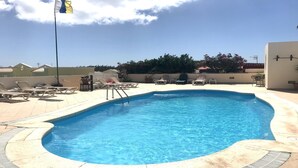 A rectangular swimming pool with a clear blue water, surrounded by a tiled deck. There are lounge chairs and a small table with a potted plant nearby.