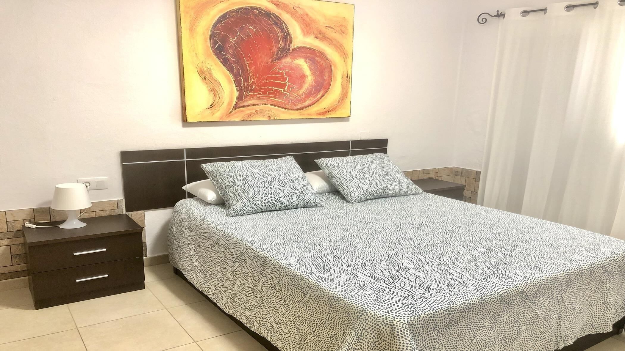 A bedroom with a bed, two pillows, a bedside table with a lamp, and a wall-mounted artwork.