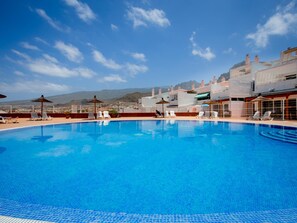 A clear blue pool with lounge chairs and umbrellas, surrounded by buildings with a mountainous backdrop.