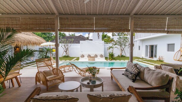 A covered outdoor seating area with wicker furniture, a round table, and a view of a swimming pool and white building in the background. - Villa M2 by Alfred in Bali (Seminyak)