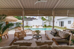 A covered outdoor seating area with wicker furniture, a round table, and a view of a swimming pool and white building in the background. - Villa M2 by Alfred in Bali (Seminyak)