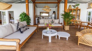 A spacious living area with a sofa, two round tables, and a wooden bench. A kitchen is visible in the background with a refrigerator and a sink. - Villa M2 by Alfred in Bali (Seminyak)