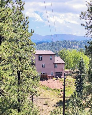A two-story house with a red roof, surrounded by dense pine trees and a mountain backdrop.