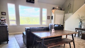 A dining area with a wooden table and benches, a black leather sofa, and a view of the outdoors through large windows. - No Cleaning Fees, Spacious & Secluded Mountain Home, Pikes Peak Views, Hot Tub! (Cripple Creek)