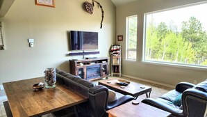 A living room with a wooden dining table, leather sofa, and a flat-screen TV mounted on the wall. - No Cleaning Fees, Spacious & Secluded Mountain Home, Pikes Peak Views, Hot Tub! (Cripple Creek)